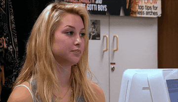 A woman with long blonde hair sits at a desk, looking to the side and chewing gum. In the background, posters and cabinets are visible