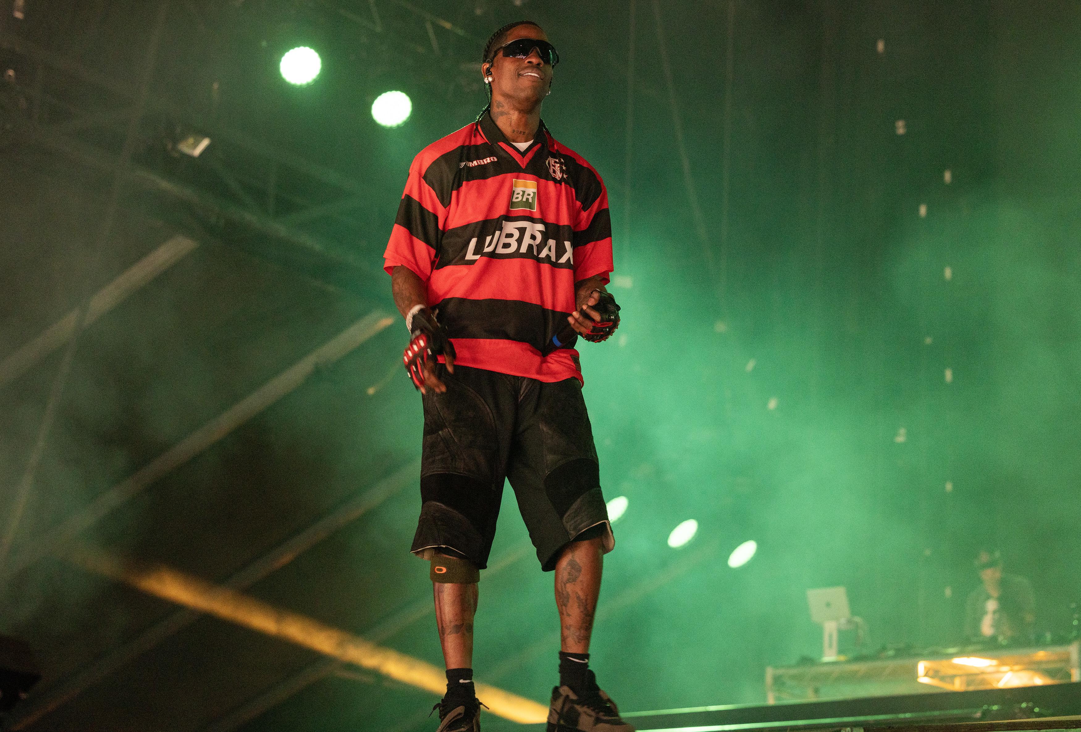 A man stands on stage wearing sunglasses, a soccer jersey, and shorts, while performing at a concert. Smoke effects and stage lights enhance the atmosphere