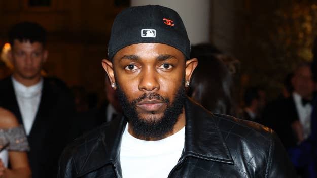 Kendrick Lamar wears a black leather jacket and a black cap at a formal event. Several people are visible in the background