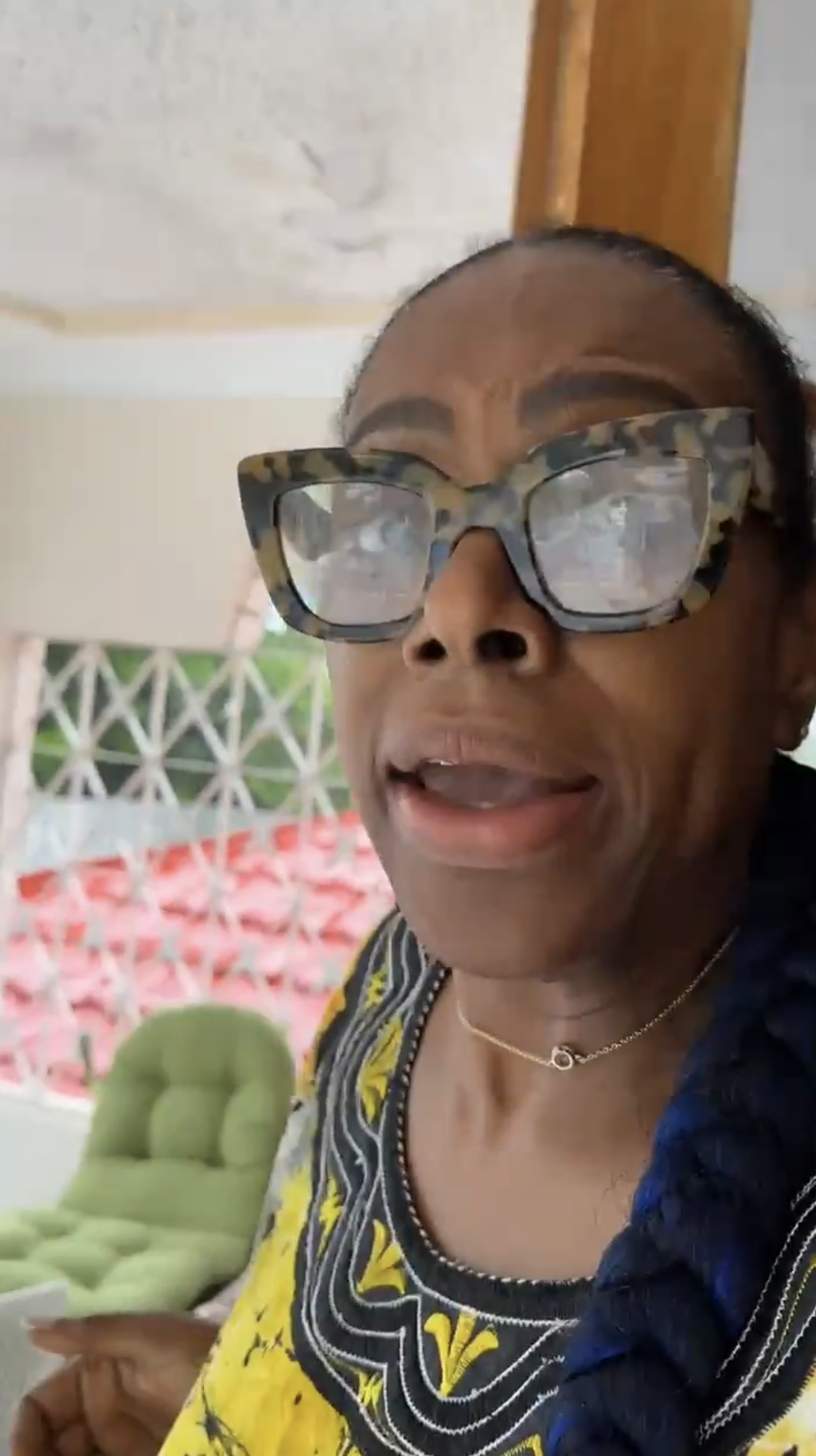 A woman with large glasses and braided hair wears a patterned top and gold necklace, speaking indoors with red and white patterned walls in the background