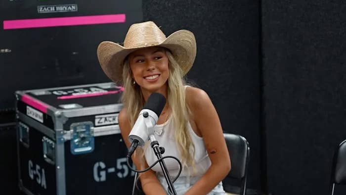 A woman in a straw cowboy hat and casual clothing smiles while sitting at a microphone during a recording session. A &quot;Zach Bryan&quot; sign is visible in the background
