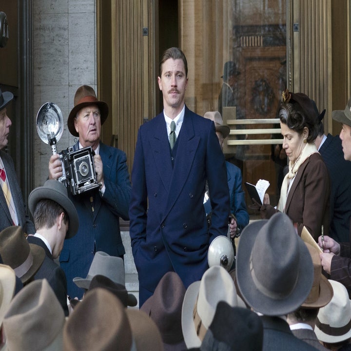 Group of people in vintage attire crowd around Garret Hedlund, standing confidently in front of a building in a scene from a film