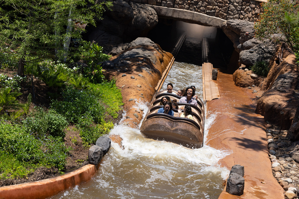Four people riding Tiana's Bayou Adventure, emerging from a tunnel into a splash. They appear excited and braced for the splash