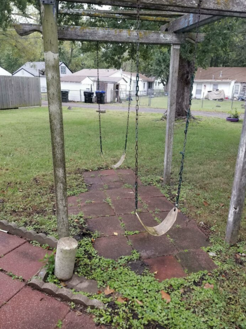 Two old swing seats hanging from chains on a wooden frame in an overgrown backyard with weathered tiles and grass