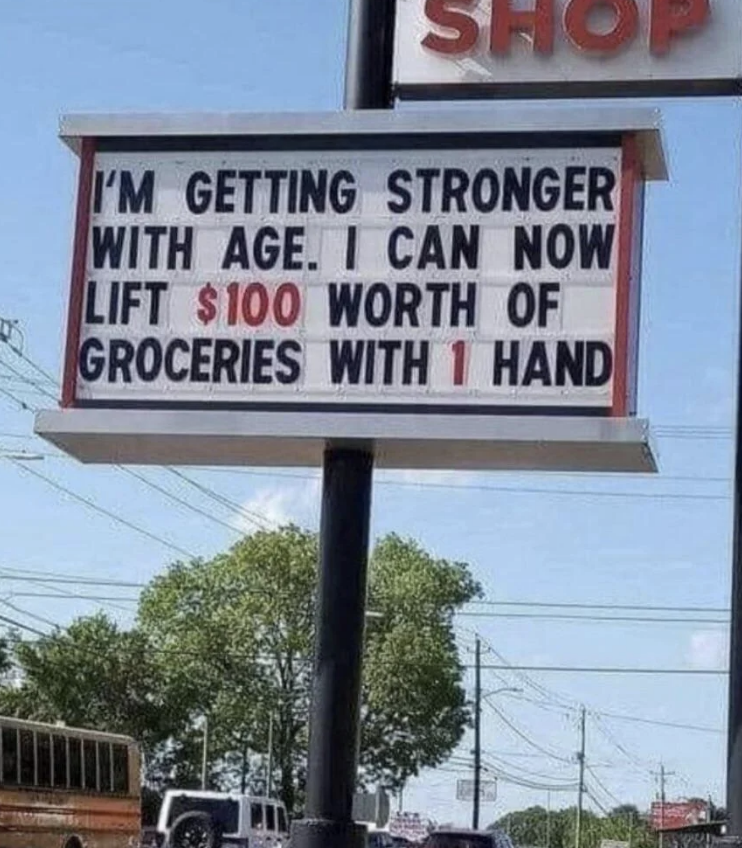 Sign reads: "I'M GETTING STRONGER WITH AGE. I CAN NOW LIFT $100 WORTH OF GROCERIES WITH 1 HAND."