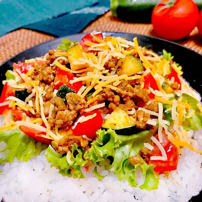 ご飯の上にレタス、挽肉、チーズ、野菜が乗ったタコライスの近接画像。背景にトマトとキュウリが見える。