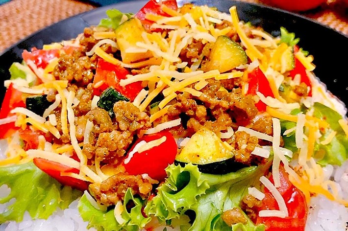 ご飯の上に挽肉、レタス、トマト、ズッキーニ、チーズが盛り付けられたタコライス。背景にはトマトとキュウリが見える。