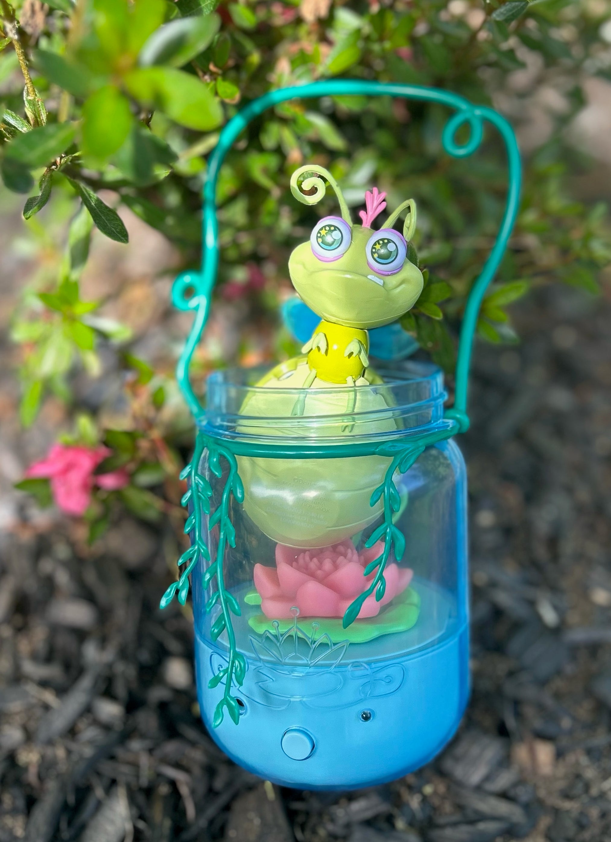 A painted mason jar lantern with a whimsical firefly figurine inside, sitting on mulch beside a bush
