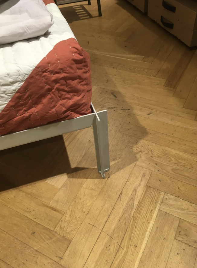 A bed in disrepair with a visibly bent and damaged bed frame leg, captured in a room with wooden flooring