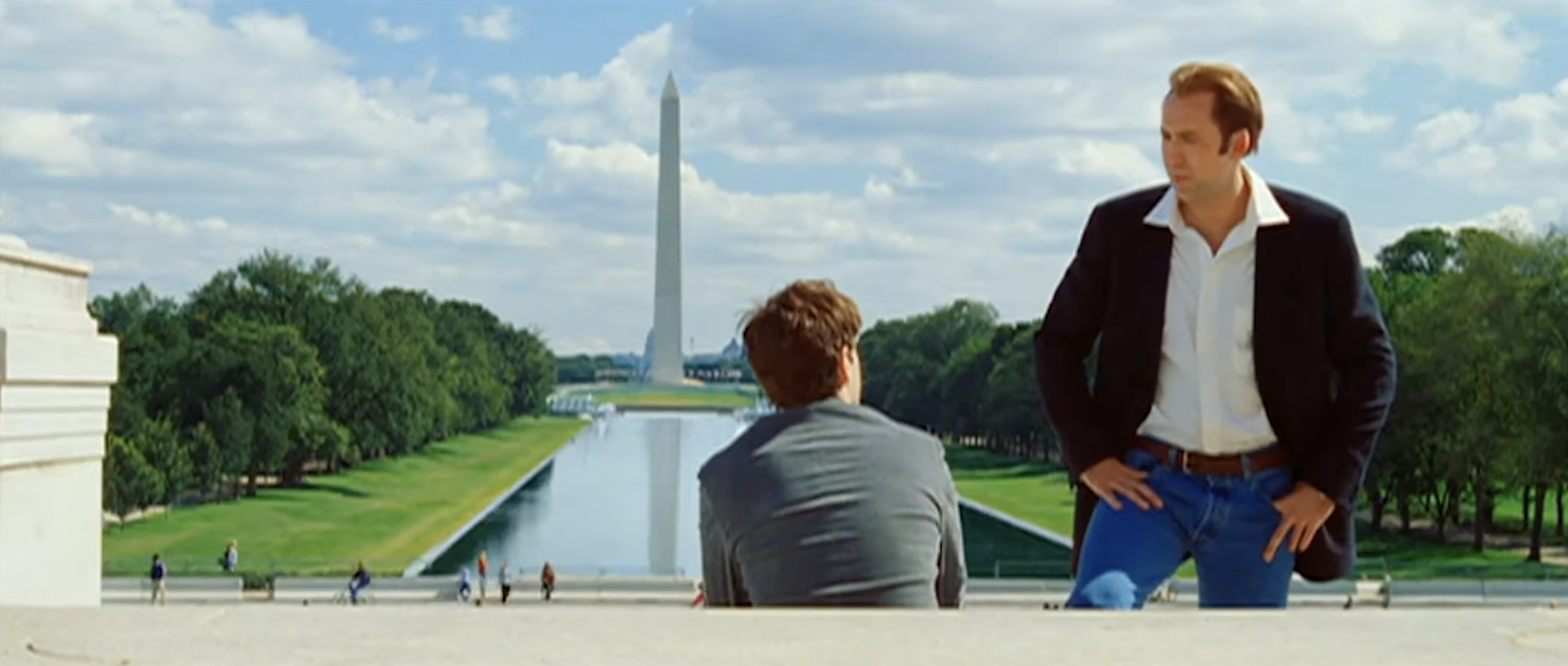Nicolas Cage and Justin Bartha in a scene outdoors, standing in front of the Lincoln Memorial Reflecting Pool, with the Washington Monument in the background