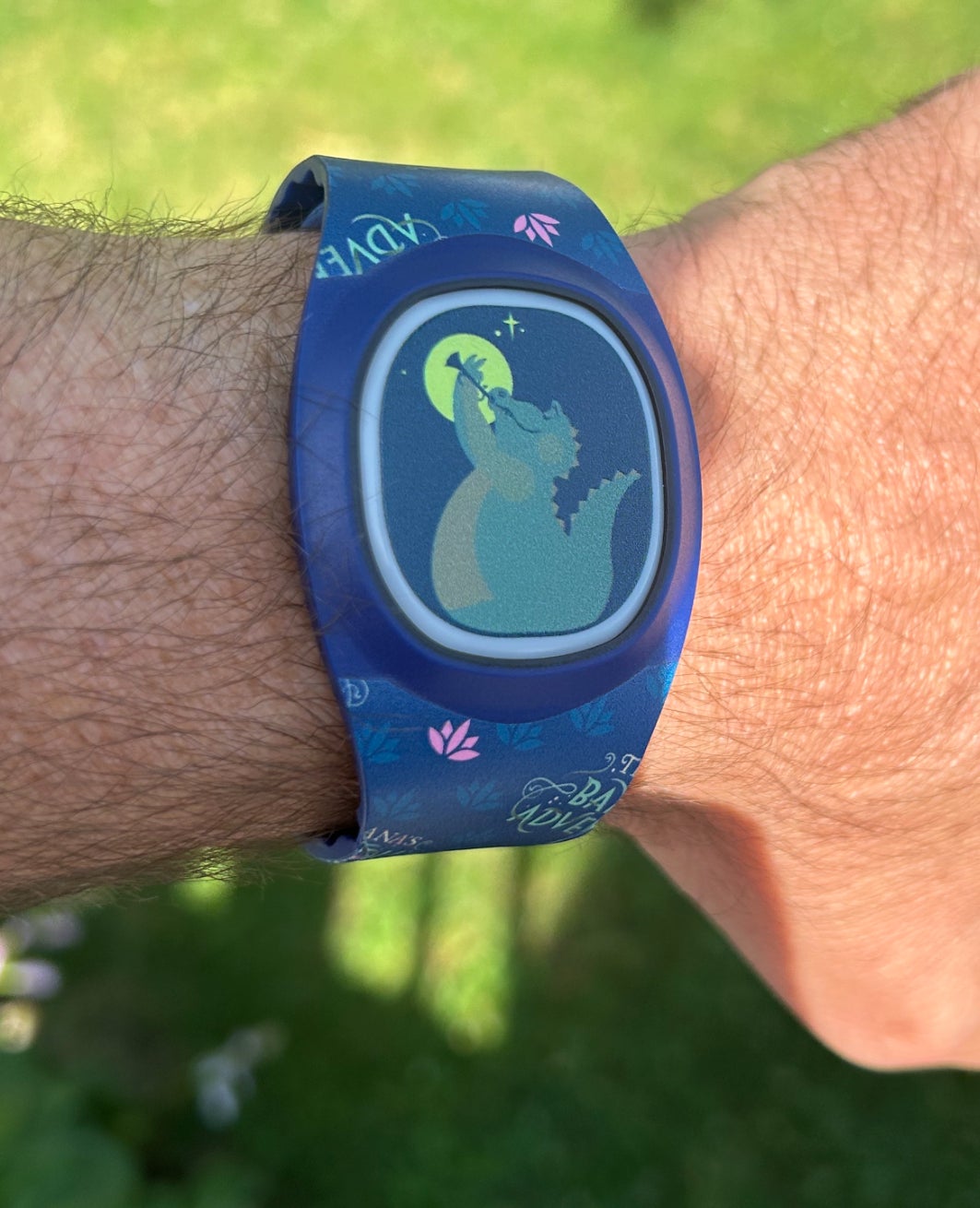 A person's wrist wearing a MagicBand+ featuring a silhouette of Louis from The Princess and the Frog in the background
