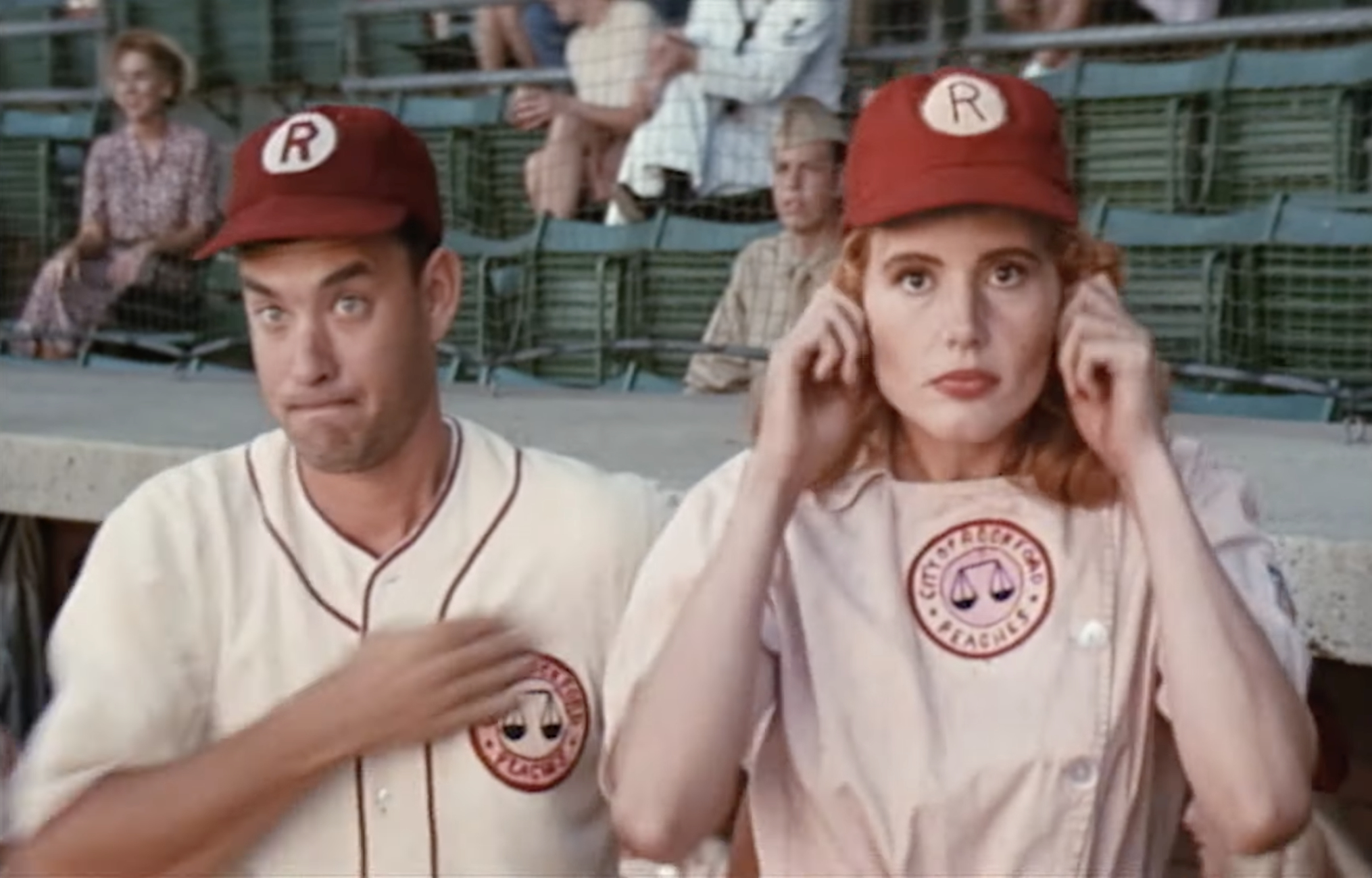 Tom Hanks and Geena Davis, in baseball uniforms, standing in a stadium; Geena covers her ears as Tom does a chest pat gesture
