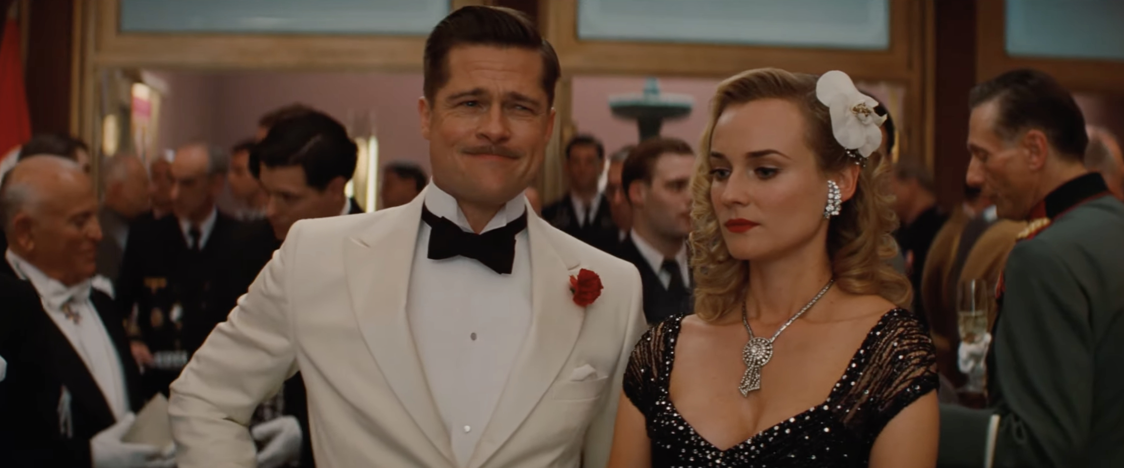 Brad Pitt wearing a white tuxedo stands next to Diane Kruger in a black, embellished gown at a formal event in a crowded room