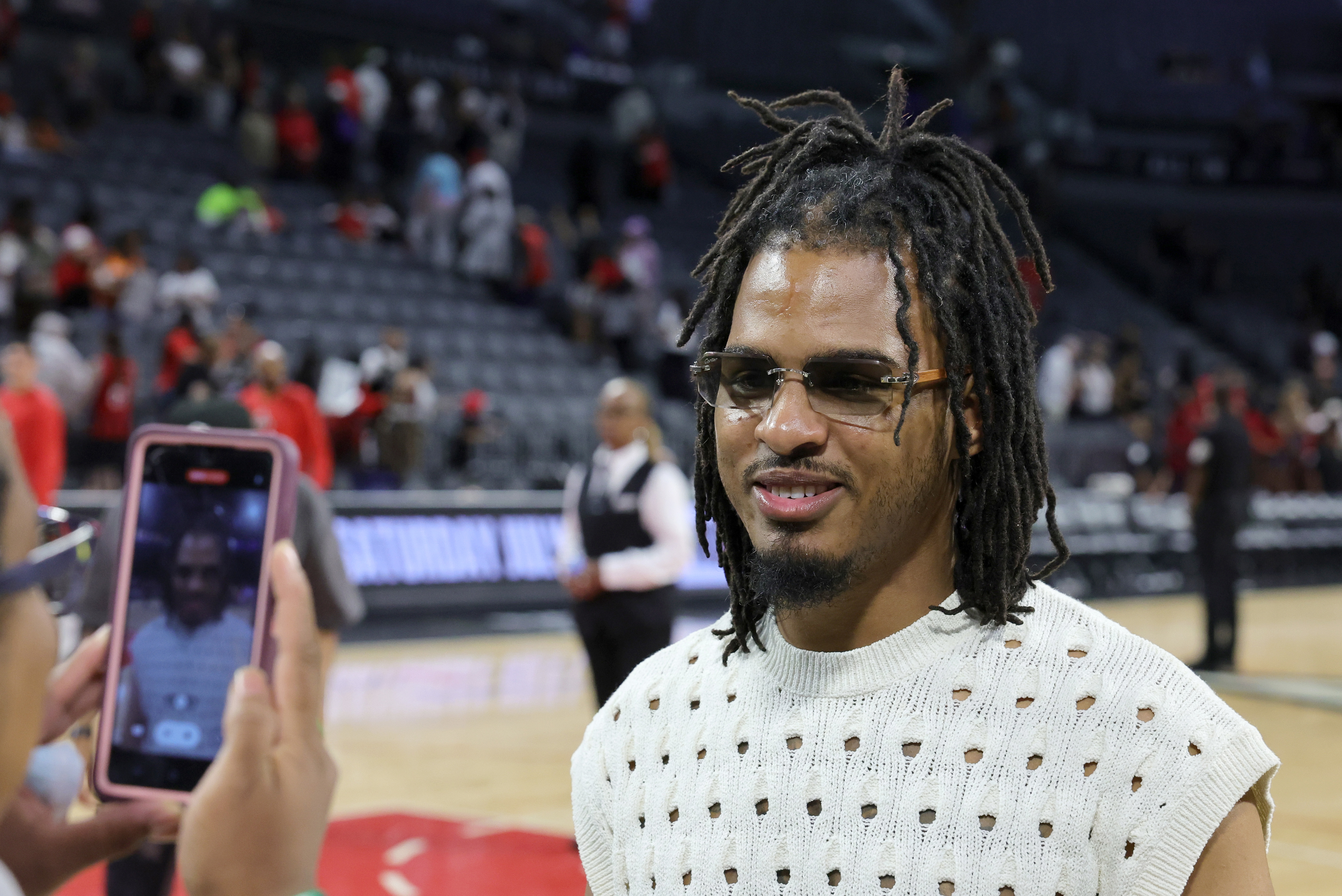 Keith Lee in a white, hole-patterned knit top and sunglasses is being photographed by another individual at a public event