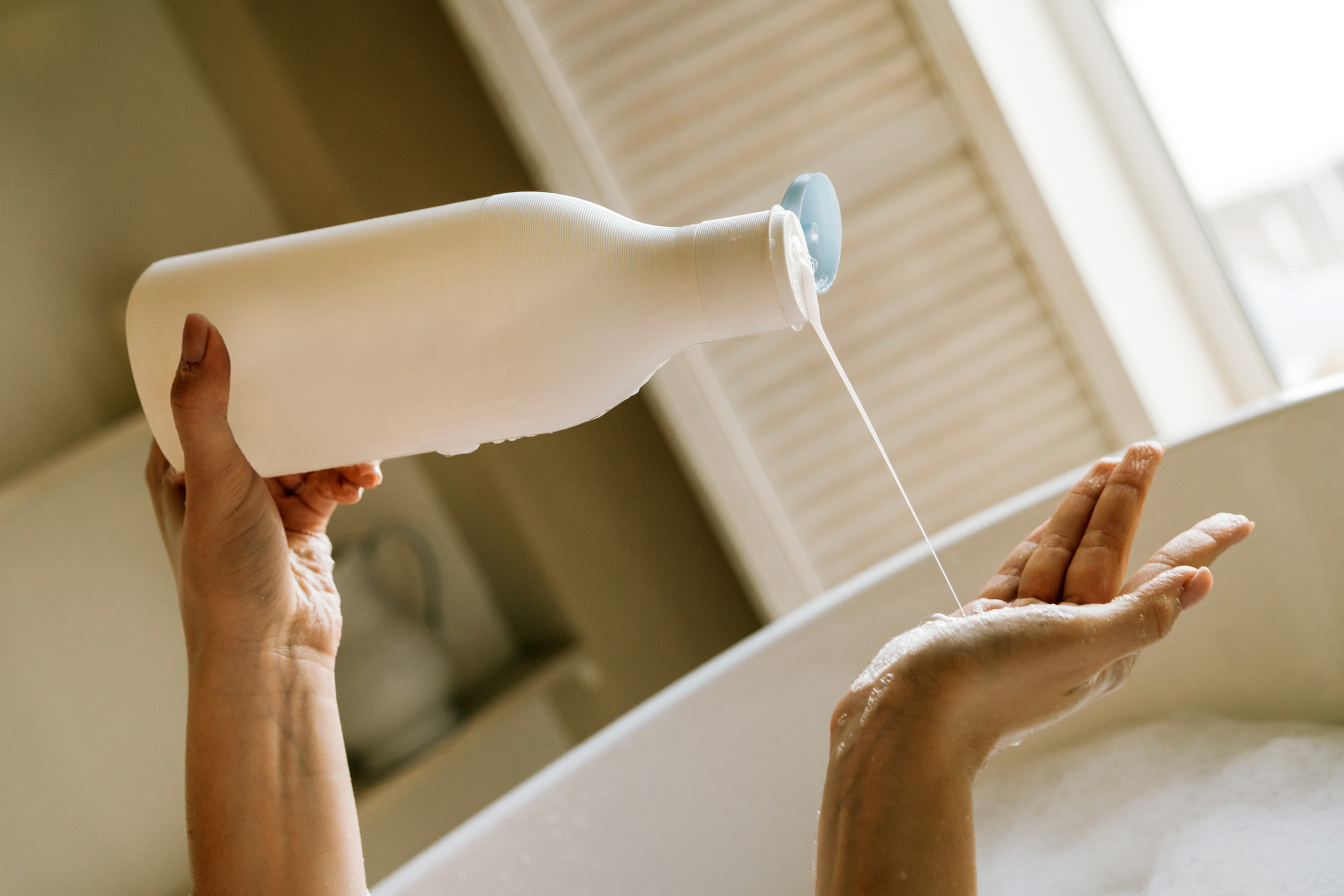 A close-up of hands, one holding a bottle of lotion, pouring lotion into the other hand over a bathtub