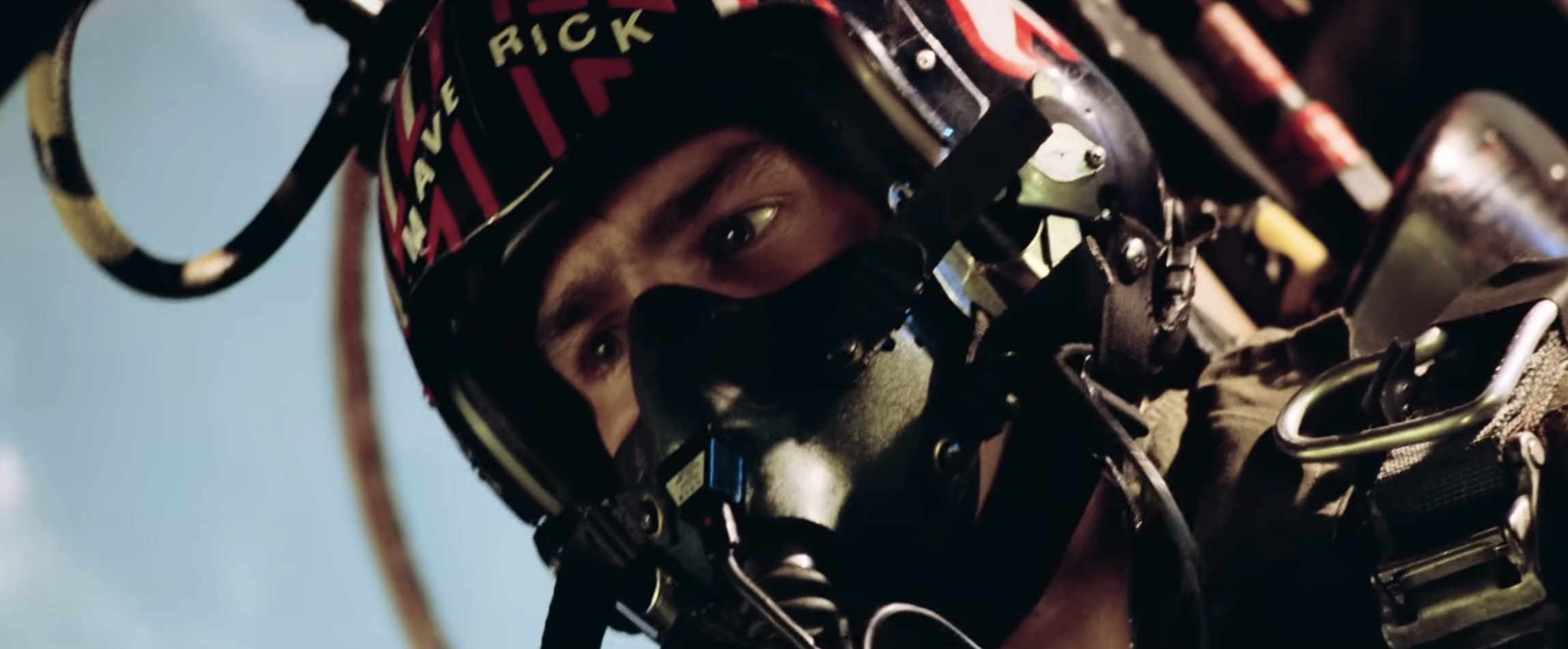 Person in a fighter pilot helmet and oxygen mask inside a cockpit, looking focused and determined. The helmet has text on it