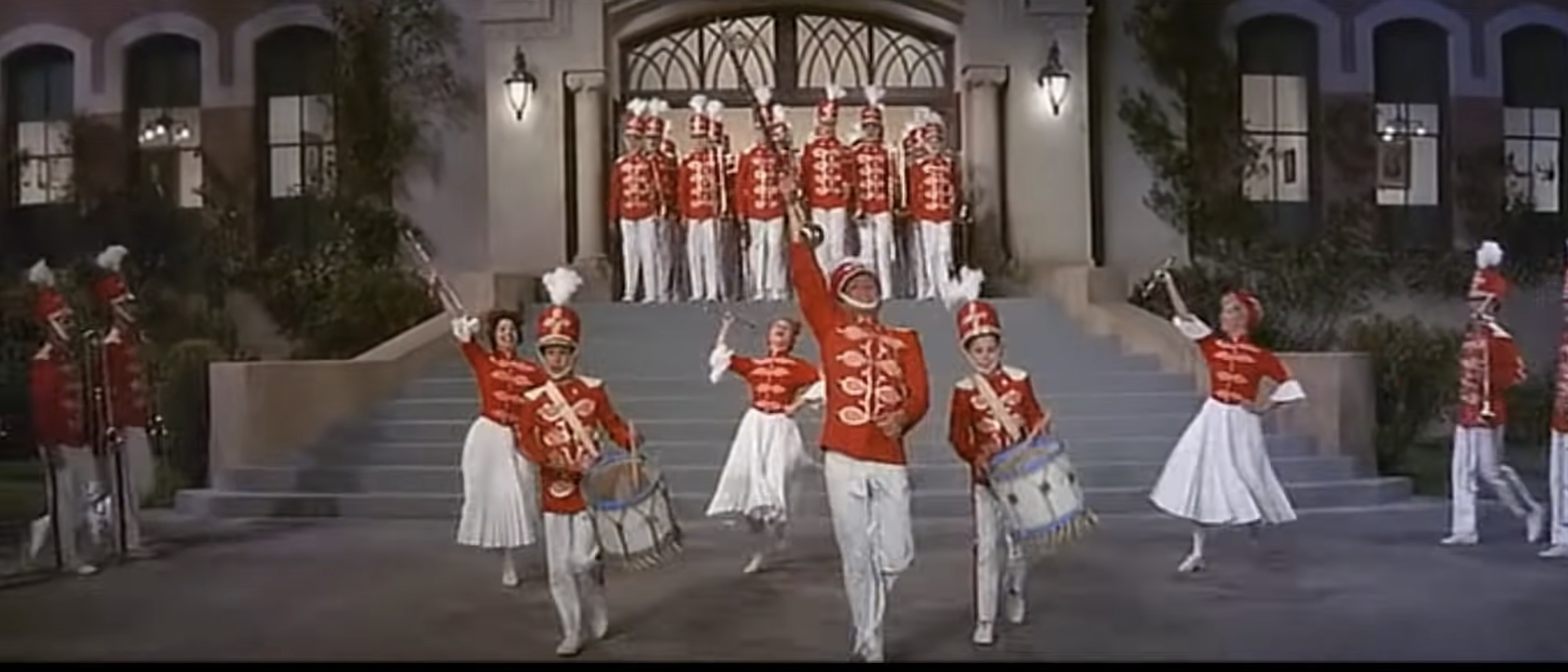 A group of performers in red and white marching band uniforms are playing instruments and dancing in front of a building with steps
