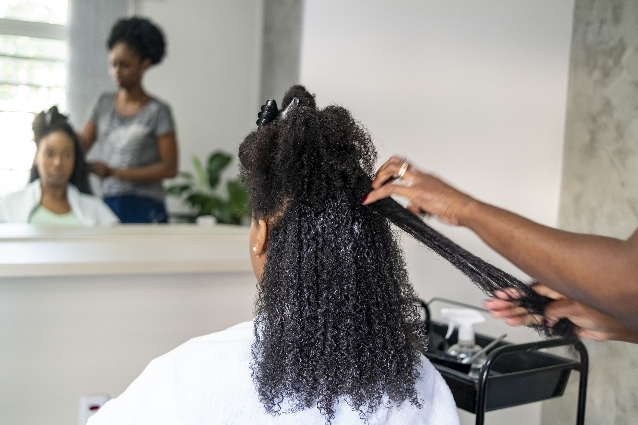 A woman styles another woman's curly hair in a salon, with a reflection of both in the mirror