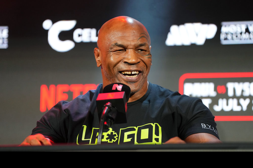 Mike Tyson at a press event, wearing a black shirt with "Lift the Game!" print and smiling while speaking into a microphone