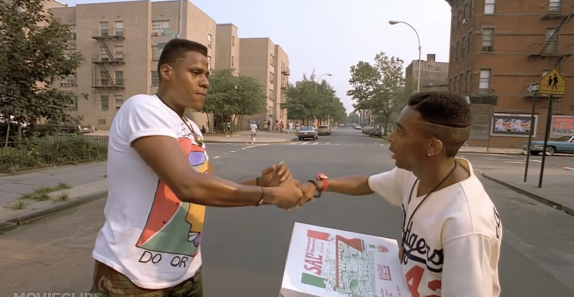 Two men shake hands on a city street. One holds a pizza box
