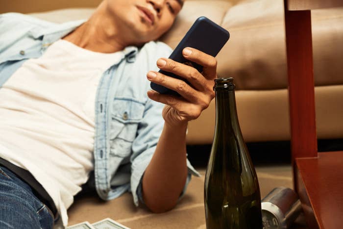 A person lies on the floor holding a smartphone, with a bottle and cans near them. Their face isn't visible, and the setting suggests a relaxed or casual environment