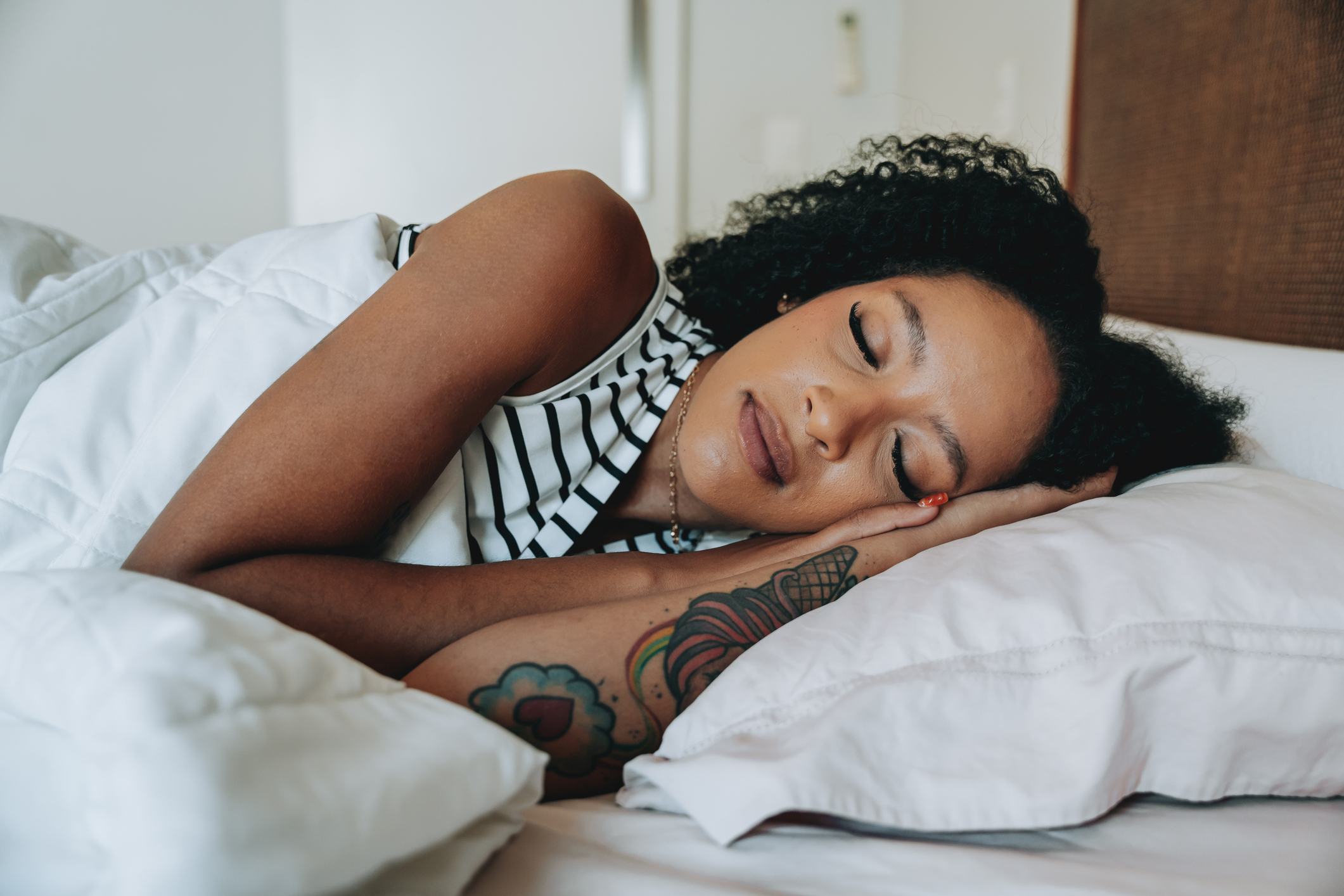 A woman with curly hair and arm tattoos sleeps peacefully on her side, with her head resting on a pillow
