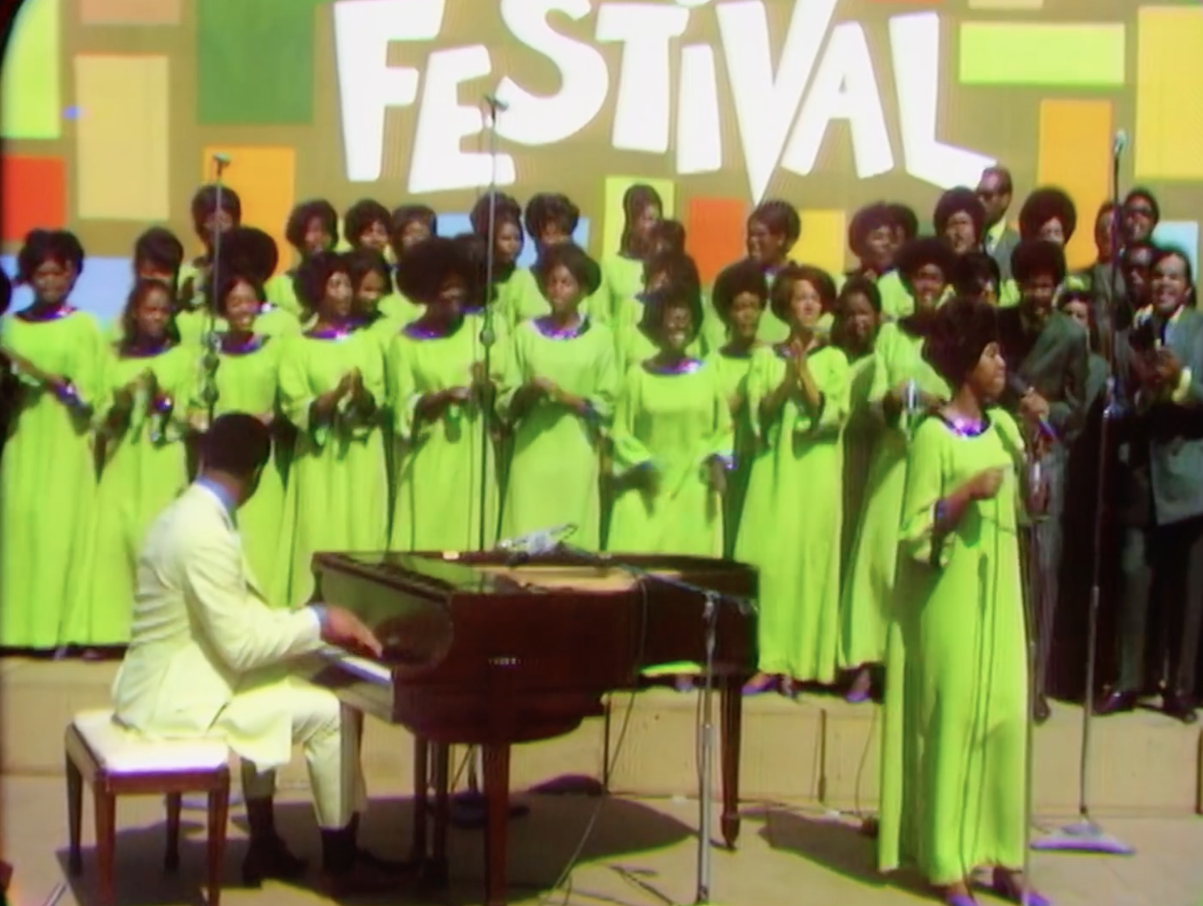 Aretha Franklin performs with a choir. The choir and Aretha wear matching outfits. An unidentified pianist plays the grand piano