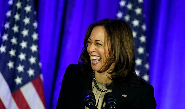 Vice President Kamala Harris laughing while speaking at an event, with two American flags in the background. She is wearing a dark blazer and a light top