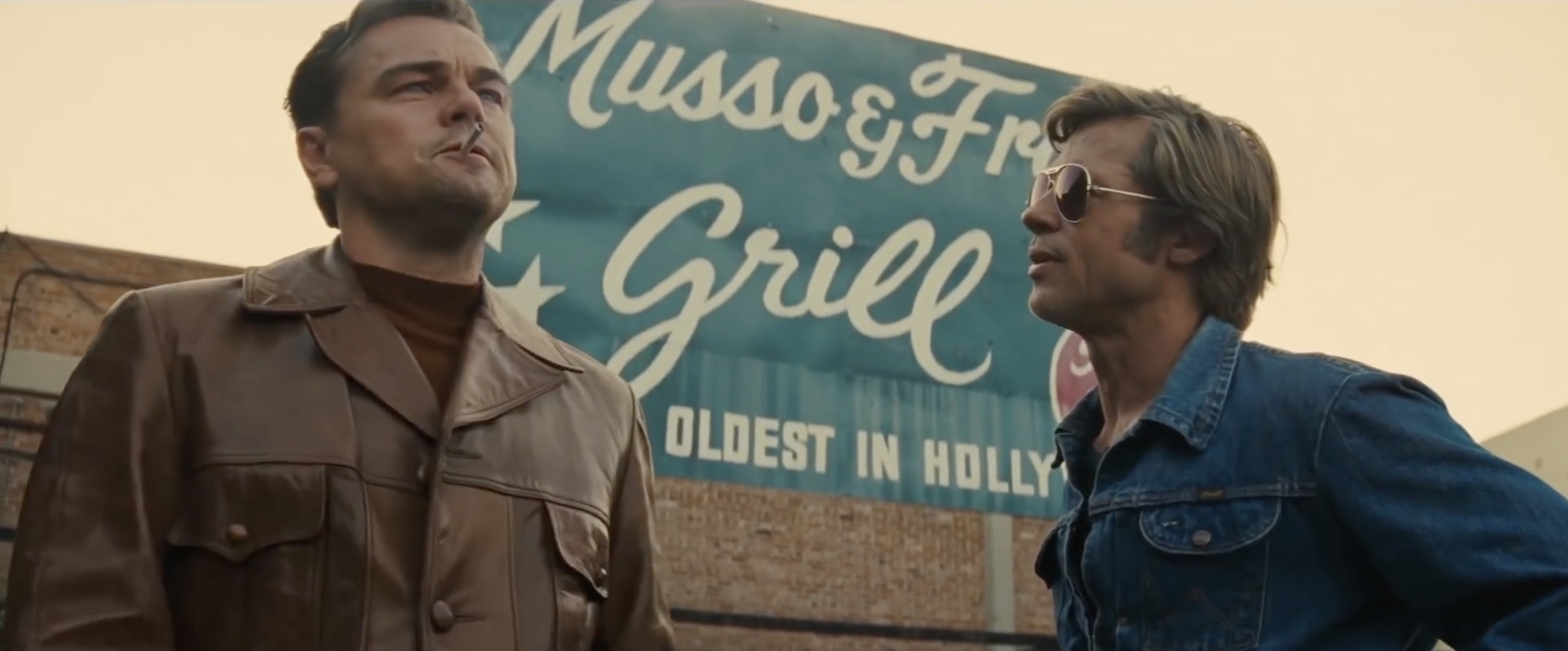 Leonardo DiCaprio in a leather jacket and Brad Pitt in a denim jacket stand outside Musso & Frank Grill