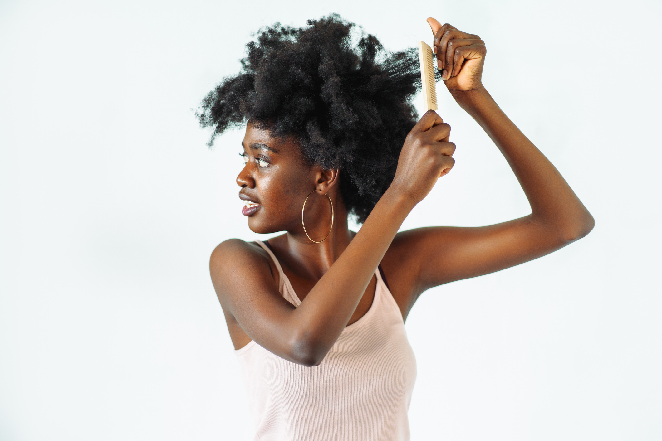 A woman combs her natural, voluminous hair while looking to her right. She is wearing a sleeveless top and hoop earrings