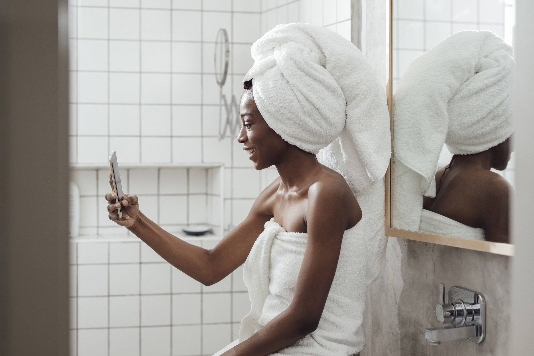 A woman wrapped in a towel with a towel on her head takes a selfie in a white-tiled bathroom