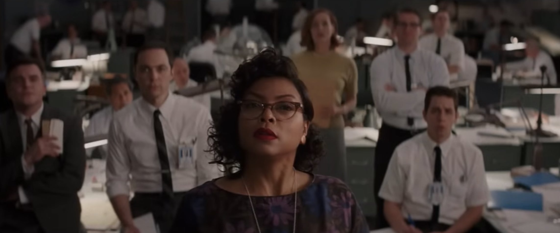 Scene from Hidden Figures. Taraji P. Henson stands confidently in office setting, surrounded by focused colleagues