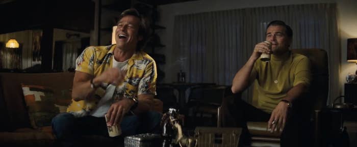 Brad Pitt, in a casual Hawaiian shirt, laughs while sitting next to Leonardo DiCaprio, who is wearing a yellow turtleneck and holding a drink, in a home setting