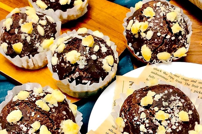 6つのチョコレートマフィンが白いマフィンカップに入っている。マフィンの上には砕いたナッツが載っている。背景に新聞と布がある。