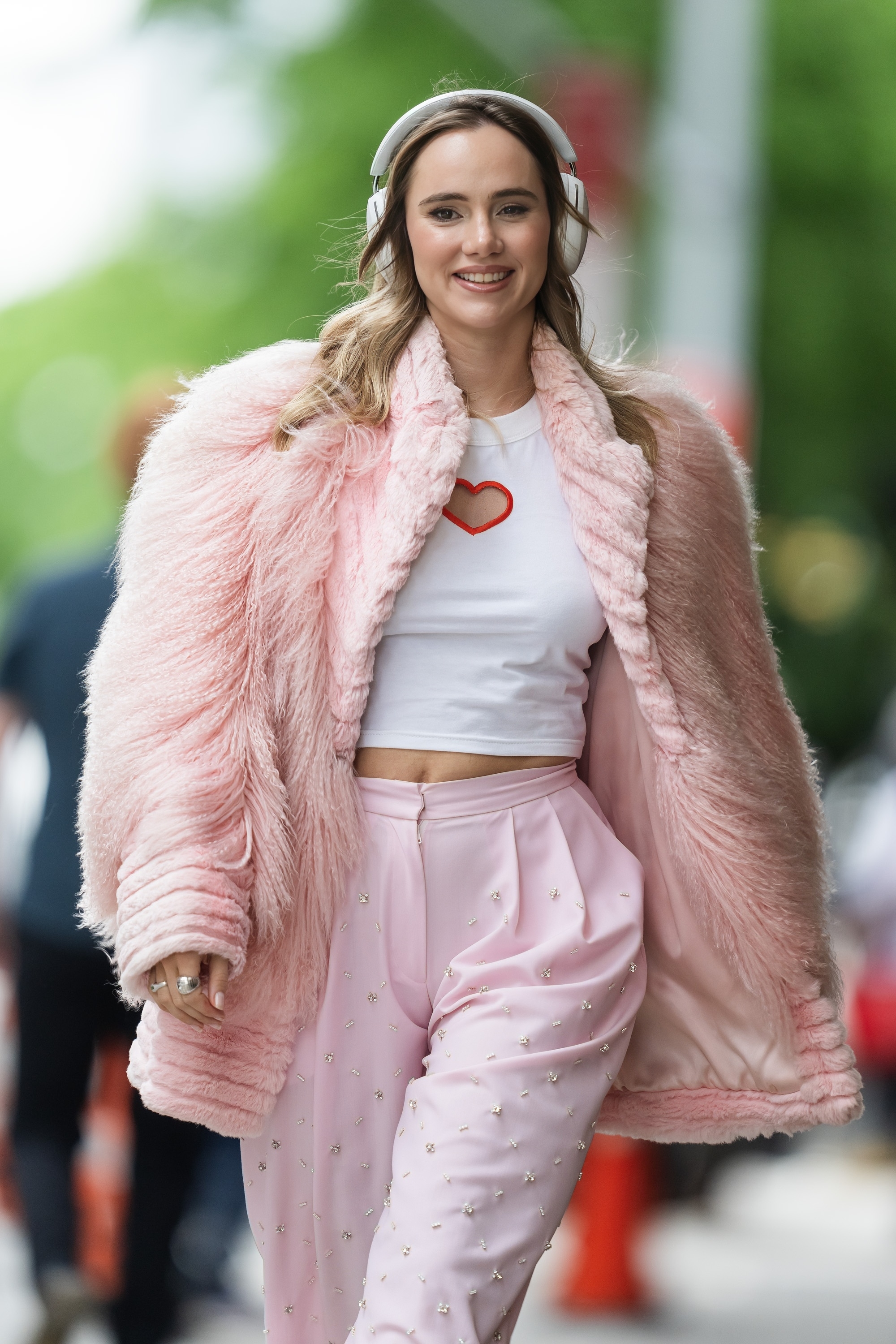 Suki wearing a fur coat, crop top with a red heart, and pants with embellishments, is walking outside while smiling and listening to headphones