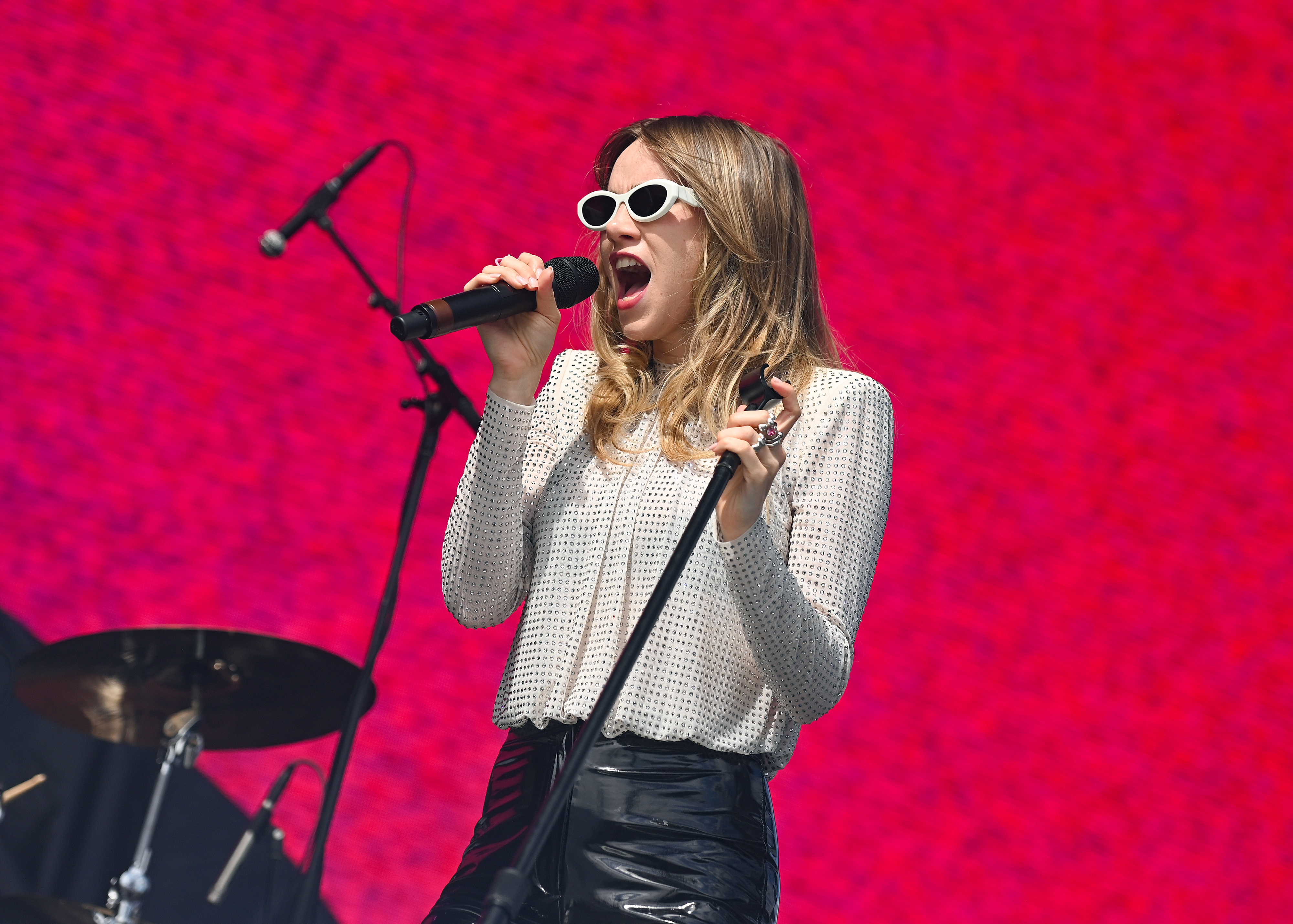 Suki in sunglasses sings passionately into a microphone on stage, wearing a long-sleeve top and black skirt. A drum set is visible in the background