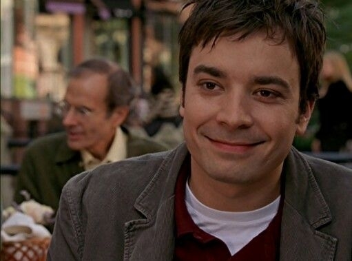 Jimmy Fallon smiles while sitting outdoors at a café. An older man is seated behind him, engaged in conversation