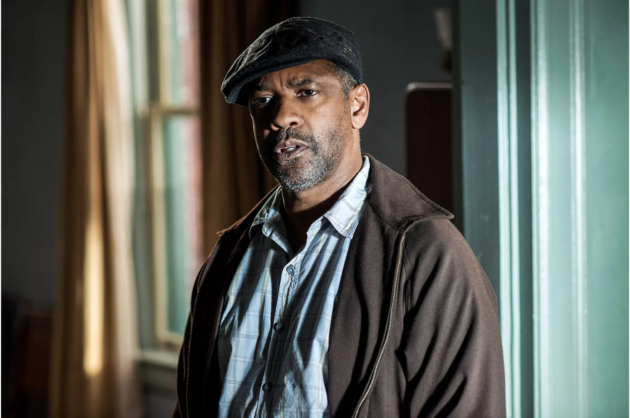 Denzel Washington wearing a flat cap and casual jacket, standing indoors with a window in the background