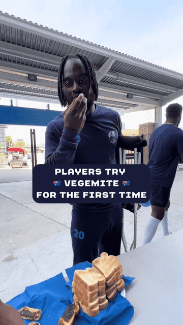 Several players try Vegemite for the first time. One player is tasting it while others are in the background. Several slices of bread are laid out on a table