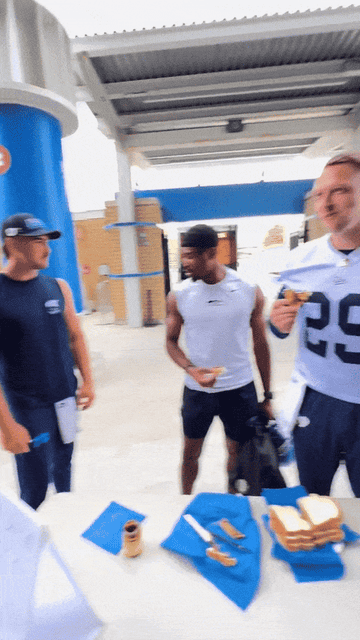 Three men, including an athlete in a white sleeveless shirt and another in a number 29 sports uniform, share a light moment while making Vegemite sandwiches