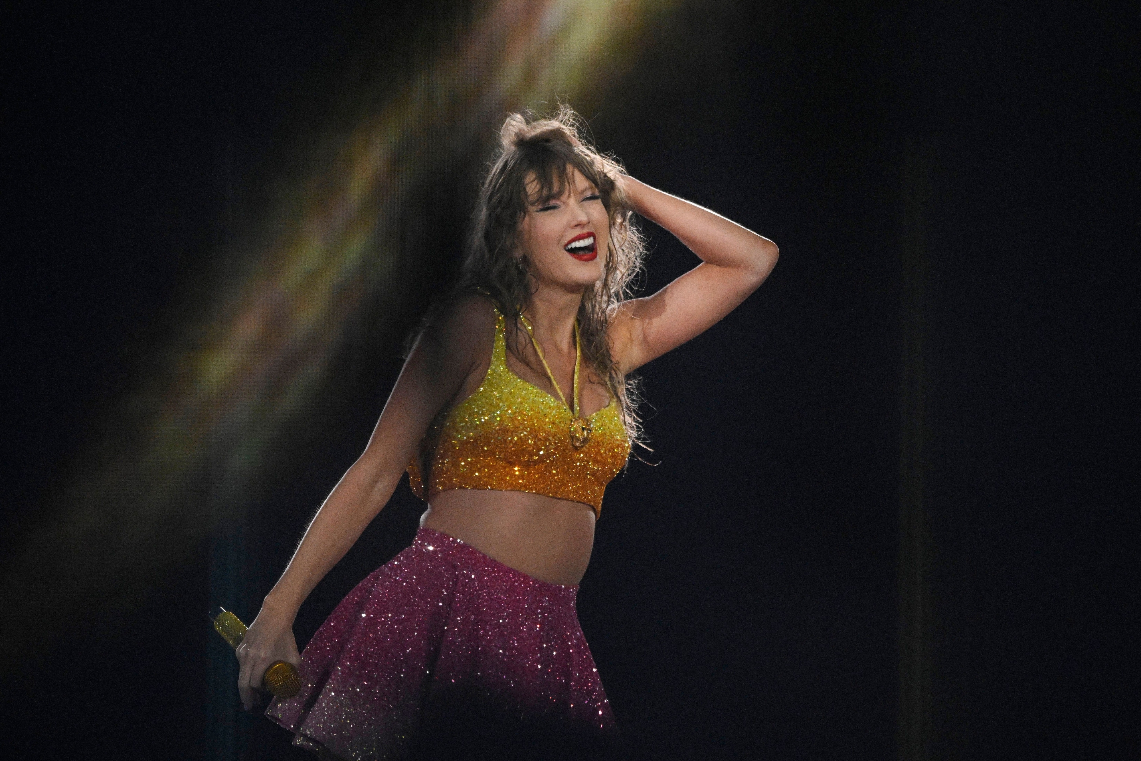 Taylor Swift performs on stage, wearing a sparkling yellow crop top and a pink skirt. She smiles warmly and holds her hair with one hand