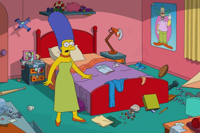 Marge Simpson stands in a messy bedroom with various items scattered around, looking surprised. A poster of Krusty the Clown is on the wall