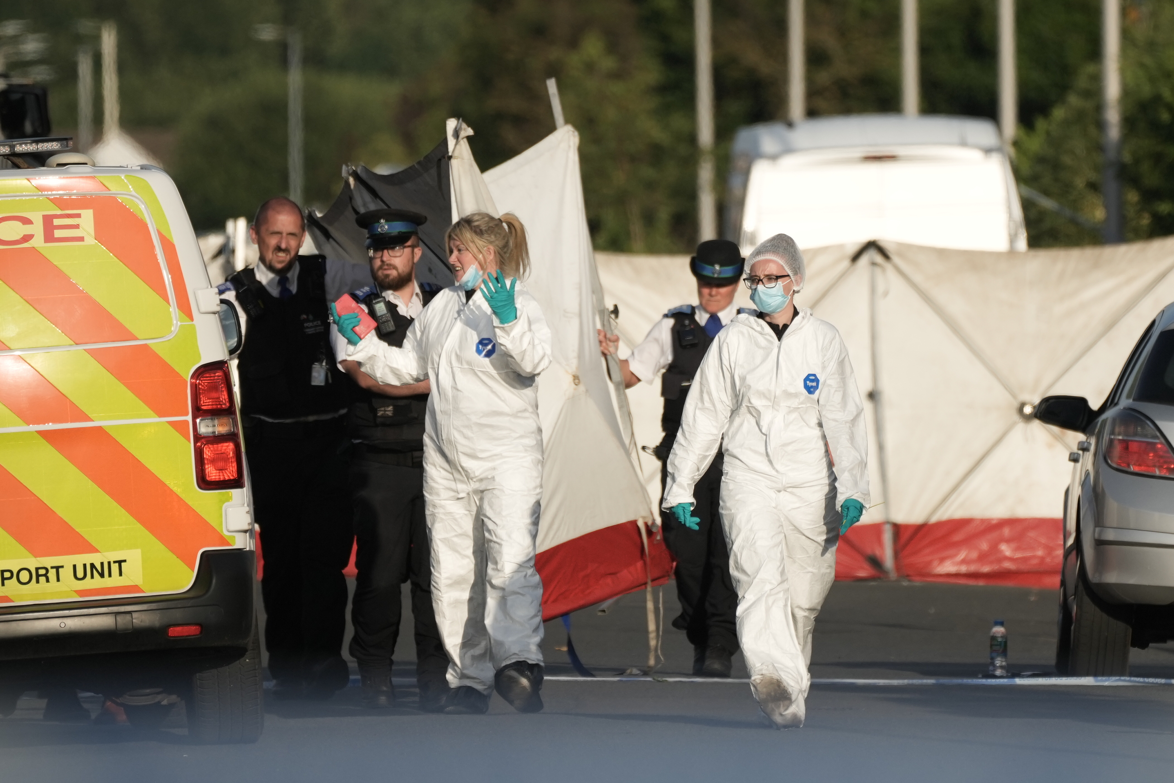 Police officers and forensic teams in protective suits work at a crime scene near several vehicles and a large white tent