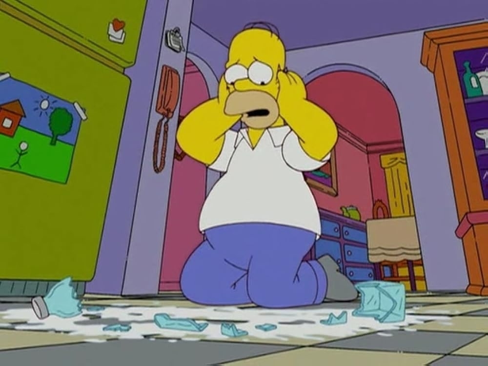 Homer Simpson kneels in distress, holding his head with both hands after dropping a broken glass jar on the floor in his kitchen
