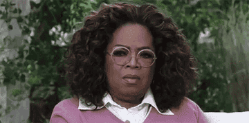 Oprah Winfrey sits outdoors, wearing a white-collared shirt under a light sweater, with a serious expression