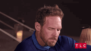 I do not know who this person is. The image shows a man in a blue shirt looking downward with a contemplative expression while seated, captioned by TLC