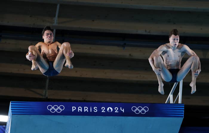Two male divers, mid-dive, performing tuck positions off a diving board with &quot;Paris 2024&quot; and Olympic rings signage in the background