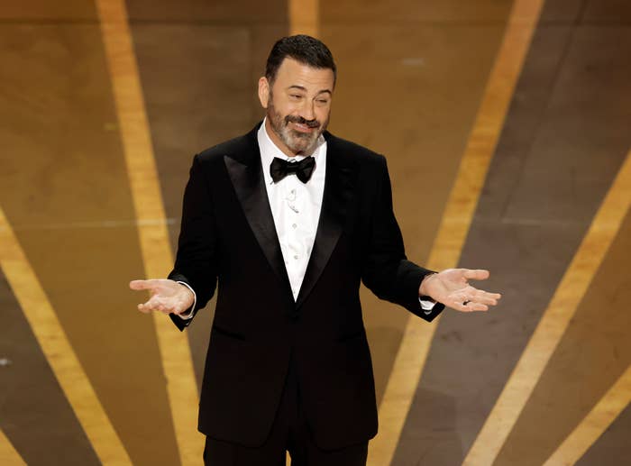 Jimmy Kimmel on stage wearing a black tuxedo with a bow tie, gesturing with both hands, likely during a speech or presentation at an event