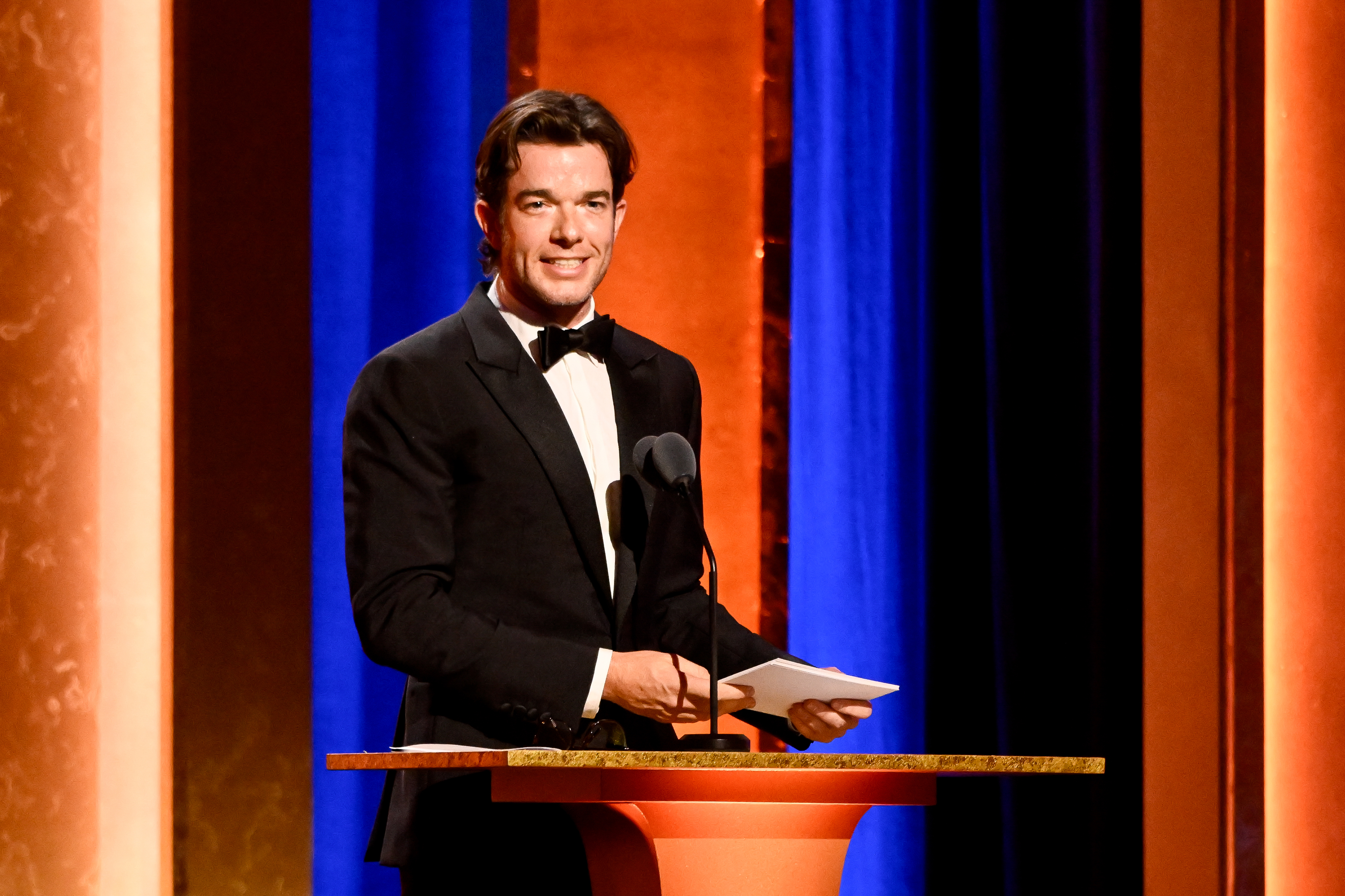 John Mulaney, in a black tuxedo, is speaking at a podium during an event