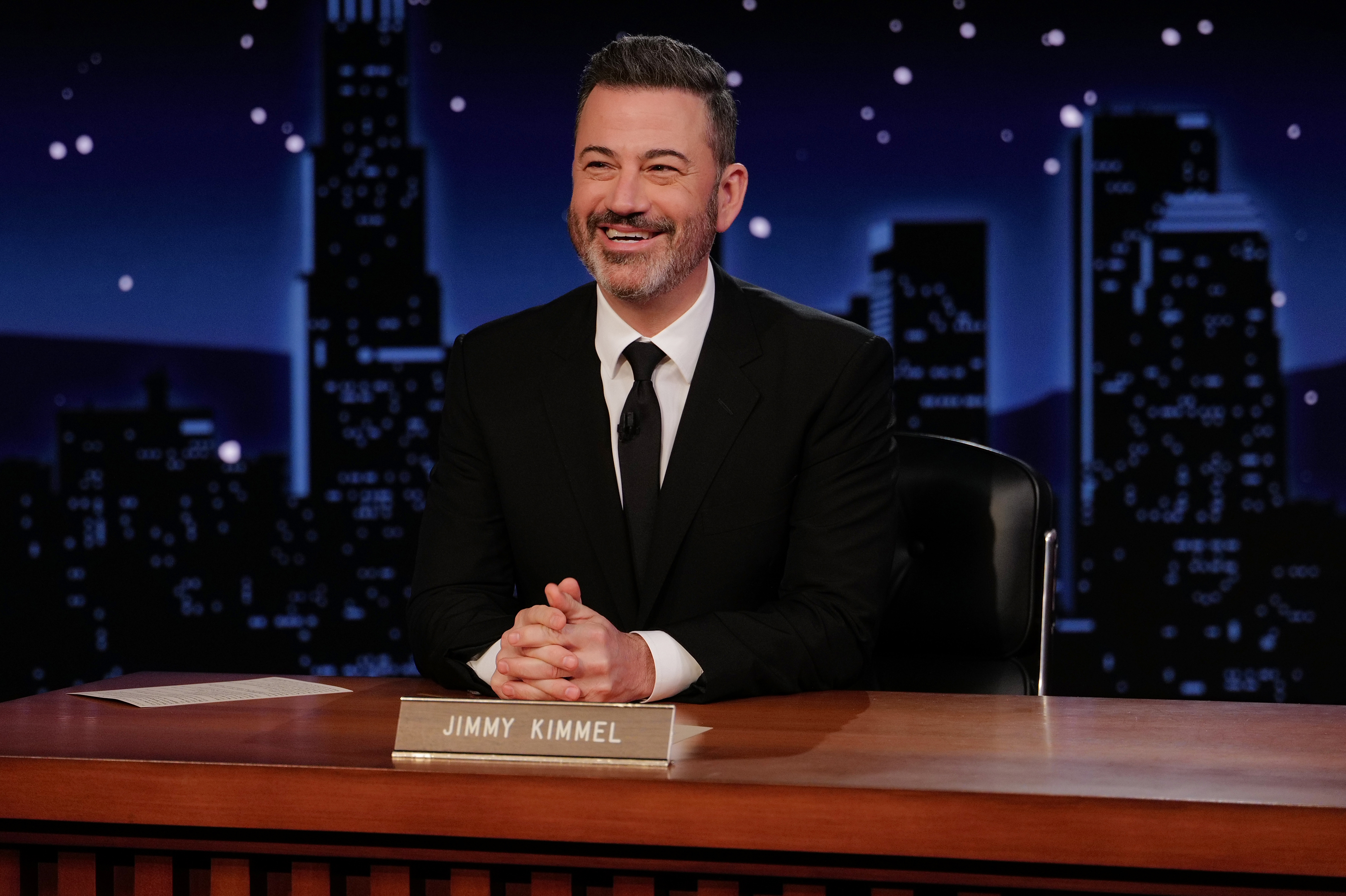Jimmy Kimmel smiling behind a desk with his nameplate on a nighttime talk show set with a city skyline backdrop